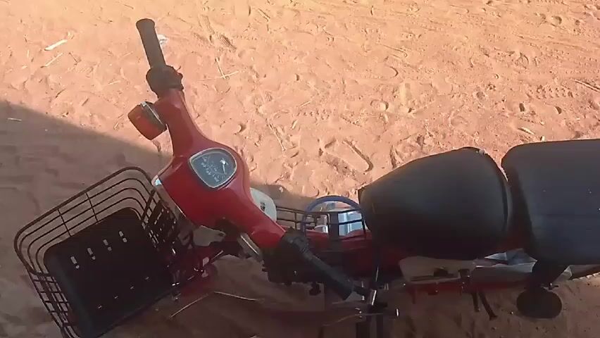 The video shows a red motorcycle lying on its side on a sandy surface. The motorcycle has a black seat, a front basket, and a speedometer. The sand is light brown and has some debris scattered around. There are tire tracks in the sand. The camera moves around the motorcycle, showing different angles.
