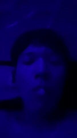 The video is a close-up, low-light shot of a person's face illuminated by blue light. The person's eyes are partially closed and their mouth is open. The person's head appears to be tilted back. The background is dark and indistinct, with some faint light streaks visible at the top.