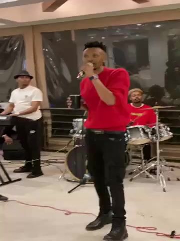 The video shows a man in a red shirt singing into a microphone, holding a cup in his left hand. He is dressed in black pants and black boots. Behind him, a man in a white t-shirt and hat plays the keyboard, and another man plays the drums. The walls of the room are white and the floor is light beige. There are large windows with curtains behind the band. The man singing is the focus of the shot.