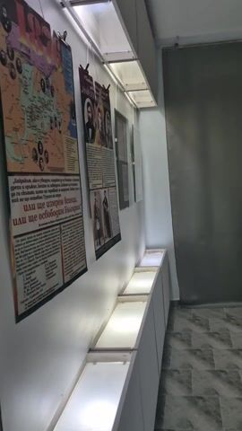 The video captures an interior view of an exhibition space, likely a museum or gallery, located in Byala Cherkva, Bulgaria. The room features white walls, a geometric patterned tiled floor in shades of gray and white, and bright artificial lighting from rectangular ceiling fixtures.

The walls are adorned with framed documents and photographs, many of which appear historical and in black and white, suggesting a historical or cultural exhibition. Below some of these wall-mounted displays are long, white, low-level display cases or shelves with integrated lighting. High on some walls are modern gray cabinets. A large section of one wall is covered with a plain gray fabric panel, and a dark, possibly glass-topped, display case is visible in the background of the initial shot.

Several individuals are seen touring the exhibition. A middle-aged woman, dressed in a gray hooded jacket, black t-shirt with a graphic, and gray sweatpants, walks through the room, looking upwards at the displays with a focused or thoughtful expression. Two young adults are also present. One young man, with long dark hair, wearing a black leather jacket over a dark hoodie, walks and offers a slight smile. Another young man, wearing glasses and a dark long-sleeved shirt, is shown standing, holding a gray garment, and looking towards the camera with a neutral expression. A young woman, with long dark hair, dressed in a black t-shirt and light-colored pants, is seen walking while holding a clear plastic water bottle and a jacket.

The overall activity depicts visitors calmly browsing and observing the exhibits within this bright, contemporary-looking exhibition space. The specific text on the framed documents is not legible from the video.