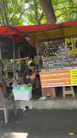 A street-side stall displays a variety of watches and watch accessories, including straps and batteries. A man is seated near the stall, possibly attending to it. The stall is set up outdoors, with trees and foliage visible in the background. The ground appears to be paved, with a concrete curb. The lighting suggests it is daytime.