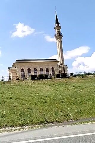 The video begins by showing a light-colored mosque with a tall minaret, situated on a grassy hill under a blue sky with white clouds. A chain-link fence runs along the top of the hill. The camera then pans to the left, revealing concrete pillars supporting an overpass. The view transitions to a highway, with cars and trucks traveling in both directions under a bright sky filled with fluffy clouds. The camera is positioned from inside a vehicle, looking forward.