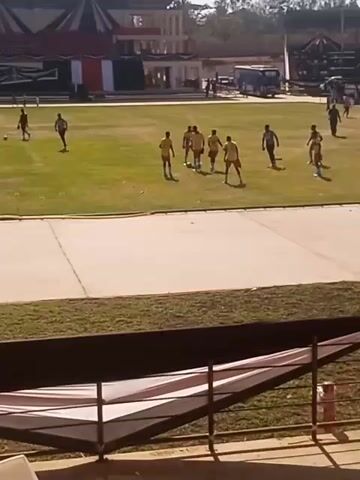 The video shows a soccer team training on a green field in an outdoor stadium. The team members are wearing yellow and dark green uniforms. They are passing and kicking a ball. Behind them, there is a building under construction. Also visible are flags and decorations in the stadium stands. Some other people are also on the field.