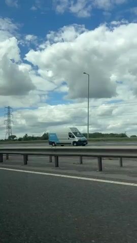 The video is shot from the perspective of a passenger inside a moving vehicle, looking out at a multi-lane highway or motorway. The weather is bright and sunny, with a vivid blue sky dotted with large, fluffy white cumulus clouds.

Numerous vehicles, including several cars (white, yellow, grey, black) and a white van, are visible driving on the road. Some vehicles are seen passing the viewer's car, while others are in the distance or on parallel lanes. The road surface is dark asphalt with clear white lane markings.

To the right of the viewer's car, a double-level metal crash barrier separates the immediate lane from the roadside. Beyond the barrier, tall, dark streetlights, utility poles carrying power lines, and a prominent large metal lattice transmission tower (pylon) stand against the sky. Green foliage, consisting of trees and bushes, lines the horizon beyond the infrastructure.

The overall scene depicts a typical daytime drive on a highway, suggesting a calm and clear day. Given the stated location context, this is consistent with a motorway in the Basildon, United Kingdom area, with traffic appearing to flow on the left.

Gentle vocal music, featuring a female singer, plays throughout the video. A snippet of lyrics audible includes "You remind me every day..." No visible text is clearly legible on any signs or vehicles.