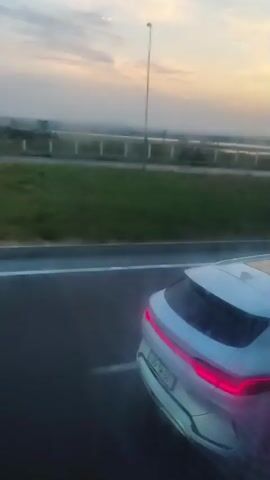 The video shows a white car driving on a highway. The sky is cloudy and the sun is setting. The car is a sedan and it has a sunroof. The car is moving at a moderate speed. There are other cars on the road, but they are not as clear. The scenery is green and there are trees in the background.