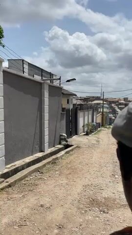 The video shows a dirt road in what appears to be a residential area. The road is lined with walls and fences. There are power lines running overhead. In the distance, there are buildings and people walking. A yellow van is parked to the right. The sky is cloudy. A drainage gutter runs along one side of the road.