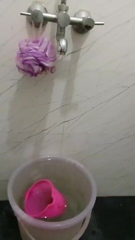 A utilitarian indoor bathroom setting is depicted in a static, medium shot. The main focus is on a metal faucet mounted on a wall of pale, simulated marble. A small stream of water flows from the faucet. To the left of the faucet hangs a fluffy, lilac-colored bath loofah. Below the faucet, a light grey plastic bucket, already containing some water and a bright pink plastic cup partially submerged within, sits on a dark surface. There is nothing else discernible in the frame.