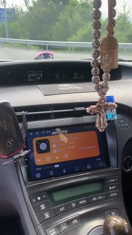 The video is filmed from the interior of a car, looking forward. The dashboard is visible, with a Toyota logo and a touchscreen display showing a music player interface. A string of wooden prayer beads with a crucifix hangs from the rearview mirror, along with a small blue air freshener. A phone mount is attached to the dashboard to the left of the screen. Outside the car, a highway is visible with several cars and two figures in orange clothing walking on the shoulder of the road. The sky is cloudy, and trees line the sides of the road. The car appears to be in motion.