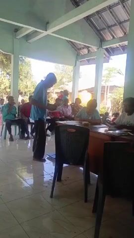 A group of people are gathered indoors, likely at a community center or meeting hall. In the foreground, a young man in a blue t-shirt and dark pants stands, holding a piece of paper and speaking to an older woman sitting behind a wooden desk. Several other people are seated in chairs behind the young man, observing the interaction. A black plastic chair is positioned between the young man and the woman. The room has a tiled floor and a light-colored ceiling with visible beams and roofing. The environment appears to be a covered outdoor space, possibly a veranda or an open-air hall, with sunlight filtering in from the side, illuminating the scene and casting shadows. Trees and buildings are visible in the background through the open sides of the structure. The overall atmosphere suggests a formal or semi-formal gathering, potentially a meeting, registration, or service point.