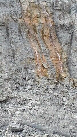 The video shows a close-up of a rugged rock face, possibly in a quarry or an excavated area. The rocks are predominantly grey, with sections featuring prominent reddish-brown and orange staining, potentially from mineral deposits. Several thin, wavy white streaks run across the reddish-brown sections, indicating paths where water is actively flowing down the rock face. To the right, there is a pile of loose, broken grey rocks. The overall scene suggests an industrial or natural environment where rock is exposed and water runoff is present. The ambient sound of machinery can be heard, supporting the idea of an active work site. No people or visible text are present. The location context provided is Hache, Nigeria.