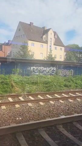 The video shows a view from a moving train looking out a window. In the foreground are train tracks. Beyond the tracks is a grassy area with some weeds and a low wall or platform. Behind the platform is a building, possibly a train station, with graffiti on its side. Further behind the building are trees and a cloudy sky. A billboard is visible on the right side of the frame with a red heart design.