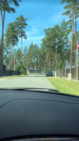The video is a first-person perspective from inside a vehicle traveling down a paved residential street lined with tall pine trees and modern houses. The sky is clear and blue with a few wispy clouds. There are fences along the sides of the properties, some with gates. A black car is parked on the right side of the street, and another vehicle is visible further down the road. There are flags on flagpoles in front of some houses. The video is shot in daylight on a sunny day.