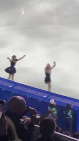 The video shows three female ice skaters in short dresses waving to an audience from an ice rink. The skaters are on a section of the rink near a low blue barrier, and the audience is visible below the barrier. The rink appears to be indoors, with a pale sky or ceiling above. The skaters are dressed in different styles of dresses, one in navy with a puffy skirt and two in black. The video is shaky and taken from a low angle.