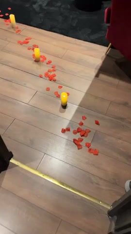 The video pans across a room decorated for a romantic occasion. Red rose petals and small yellow LED candles are scattered on a wooden floor, leading towards a large bed covered in more rose petals. To the left, a large black bathtub is filled with balloons of various pastel colors. Further into the room, a massage table with a red surface is visible. The walls are dark, and the lighting is dim, creating an intimate atmosphere. The video ends by showing a projector screen on the right side of the room. There are no people visible in the video.