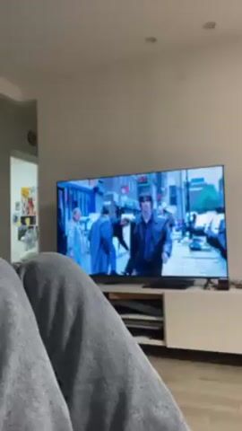 A person wearing gray sweatpants is watching television in a living room. On the TV, a scene of a man in a black leather jacket walking down a city street can be seen. Other pedestrians and parked cars are visible in the background.  The TV is sitting on a white entertainment center. There is a doorway to another room to the left. The walls are white.