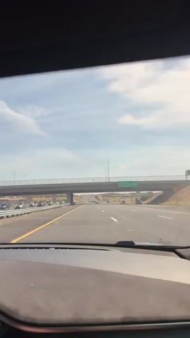 The video shows a first-person view of driving on a highway. A concrete overpass with a metal guardrail is visible overhead, with cars passing underneath. The highway has multiple lanes and is surrounded by dry, arid land with some sparse vegetation. The sky is blue with scattered white clouds. The car dashboard is partially visible in the foreground. The overall scene depicts a typical highway driving experience on a clear day.