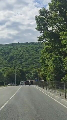 The video shows a car driving on a two-lane road toward a hill covered in lush green trees. The sky is cloudy with patches of blue. There are cars in front of the camera car, and street lights line the side of the road. A building is visible on the right side of the road with a speed limit sign of 40. Music is playing in the background.
