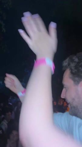 The video shows a group of people at an outdoor event at night. The initial shot focuses on a person's wrist with a pink bracelet, then pans to show a crowd under a structure with blue and yellow lights. It then briefly shows a close-up of a person's face before returning to the crowd, where individuals are seen interacting and enjoying the music. The scene is dark, with the lighting emphasizing the crowd and the event's atmosphere.