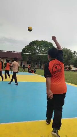 The video shows an outdoor volleyball game. There are several young people on a blue and yellow court. A volleyball is in the air. One person is serving or hitting the ball, and others are positioned to receive it. In the background, there is a building with a red roof, trees, and a cloudy sky. The players are wearing orange shirts and dark pants. The game appears to be in progress.