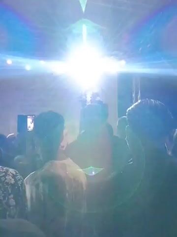 A performer sings on a stage, backed by a banner with text and lights. A crowd watches from below, many holding up their phones to record. The stage is lit with overhead lights, and the background is dark, suggesting it's nighttime. The performer is a woman with blonde hair, wearing a dark outfit with elaborate trim. The banner behind her has text in a language that appears to be Cyrillic. The crowd is diverse in age and gender, with most of them facing the stage. The overall atmosphere suggests an outdoor concert or festival event.