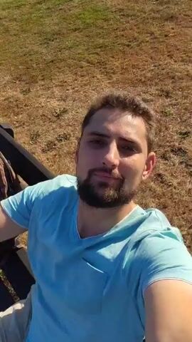 A man with short brown hair and a beard wearing a light blue t-shirt sits on a black park bench. He is taking a selfie. Behind him, to the left, is a brown backpack. To the right of the man, a bicycle is visible. The background is a grassy area with a tree in the distance. The lighting suggests it is daytime.