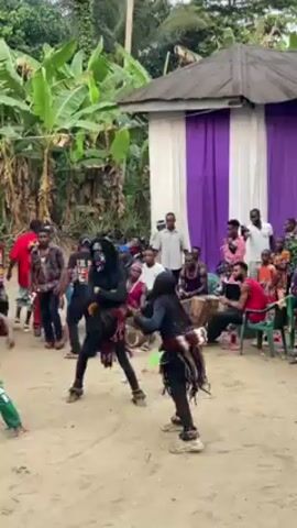 The video shows a group of people gathered in an outdoor area, observing a performance. Two individuals, dressed in matching black outfits and masks with contrasting white face paint, are engaged in a choreographed performance that looks like a fight.  A third performer, clad in a bright yellow tank top and green pants, moves in a similar style, but is further away. A crowd surrounds the performers, with some seated and others standing.  A drummer sits with the crowd, providing the background music. The setting appears to be a village or rural area, with vegetation and a building with purple and white curtains in the background. The ground is sandy.