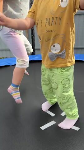 Two children are playing on a trampoline. The child on the left is wearing a yellow t-shirt with a teddy bear graphic and green pants with dinosaur prints. The child on the right is wearing a light blue t-shirt with colorful patterns and white and pink striped shorts. Both children are wearing socks. There are white tape markings on the trampoline surface, forming a cross shape. The children are jumping on the trampoline, sometimes landing on the tape markings. The background shows a metal fence and a blue trampoline safety net.