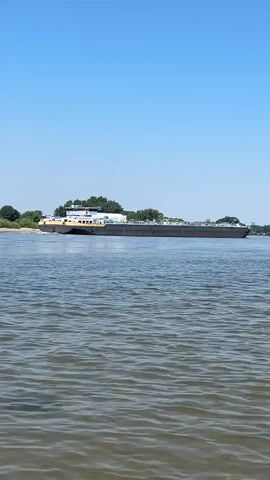 A long, flat barge with a cabin on the stern is seen on a wide river under a clear blue sky. The barge is dark gray and appears to be loaded, with a lighter colored cabin structure at the rear. The water in the foreground is rippled and brown, while the water where the barge is located is a calmer, darker blue. Trees and a sandy bank are visible in the distance behind the barge. The camera is positioned low to the water, giving the impression of being on another, smaller boat or very close to the water's surface. The barge is moving from left to right across the frame.