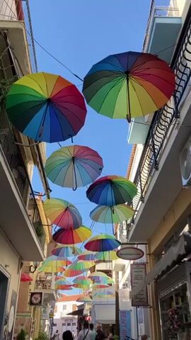 A vertical shot captures a narrow European street with buildings on either side, adorned with dozens of colorful, open umbrellas suspended overhead. The umbrellas, in rainbow hues of red, orange, yellow, green, blue, and purple, form a vibrant canopy against a clear blue sky. Sunlight filters through the gaps between the umbrellas, casting dappled light on the street below. On the ground, several people are visible, appearing to walk along the street, their faces obscured by the distance and angle. The buildings flanking the street feature balconies with railings and windows, suggesting residential or commercial spaces. Signage is visible on the buildings, hinting at shops or businesses. The overall scene evokes a festive and cheerful atmosphere, likely a tourist destination or a street decorated for an event.