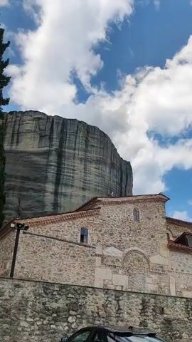 The video opens with a view of a stone building with a red-tiled roof, likely a monastery, set against a backdrop of imposing rock formations. A tall, dark cliff dominates the left side of the frame, its sheer face illuminated by sunlight. The sky is a vibrant blue, dotted with fluffy white clouds, suggesting fair weather.

As the video progresses, the camera pans to reveal more of the rock formations, which are characteristic of Meteora, Greece. These natural pillars rise dramatically from the landscape, topped with trees and ancient structures. In the foreground, the monastery's detailed stonework and arched doorways are visible. Two cars are parked in front of the building, a black sedan and a white SUV.

Further panning reveals a wider view of the area. A Greek flag and a yellow flag fly from poles on a stone walkway. The camera then shifts to capture a panoramic vista of the town of Kalabaka below, with its tiled roofs and scattered buildings nestled at the foot of the mountains. The scene evokes a sense of peace and historical significance, typical of religious sites in natural wonders. The time of day appears to be midday, given the bright sunlight and the clear sky.
