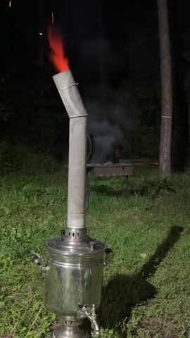 A samovar is on the grass in front of trees at night. Smoke and flames are coming out of the top of the samovar. A barbecue grill with food on it is behind the samovar.