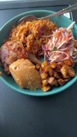A close-up shot displays a turquoise plate filled with a meal, presented against a dark, textured surface. The plate contains a serving of Jollof rice, a piece of chicken covered in a reddish sauce, a block of what appears to be bread or a similar food item, a pile of fried plantains, and a side of coleslaw. A spoon rests on top of the Jollof rice. The camera angle provides a direct, overhead view of the meal.