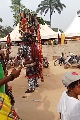 The video shows a costumed figure participating in a cultural performance. The figure is dressed in a black and white patterned outfit with a red and black striped garment over it. They also have a large structure on their head adorned with figures, plants, and other decorative elements. Several people are observing and recording the performance with their phones. The setting appears to be an outdoor event with tents, palm trees, and motorbikes visible in the background. Some of the observers are wearing traditional Nigerian attire.