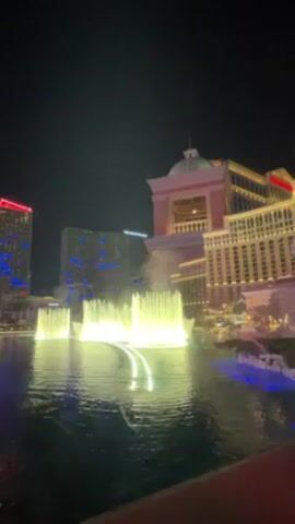 The video shows a nighttime view of the Bellagio fountains in Las Vegas.  The fountains are illuminated with bright white lights and are spraying water high into the air. In the background, several hotel buildings are visible, illuminated with various colors. The water in the fountain's pool is dark, reflecting the lights of the surrounding buildings.  The sky is dark and clear. The camera seems to be stationary.