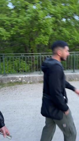 The video shows two young men walking outdoors. Initially, the focus is on the first man, wearing glasses and a black puffer jacket, as he walks alongside a low stone wall and a green metal fence, with lush green trees in the background. The camera then shifts to the second man, wearing a black hooded jacket and a crossbody bag. The first man then abruptly stops, faces the camera, and makes a gesture with his arms, obscuring his face momentarily. Afterwards, the camera resumes its movement, following the two men as they continue to walk together.