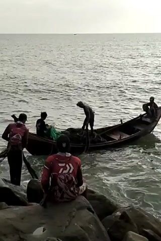 The video shows a group of five people on a rocky shoreline next to the ocean. Four people are near a small wooden boat that is partially in the water. Three of the people are on the boat and appear to be handling fishing nets. One of the people on the shore is holding a pole or oar. The fifth person is sitting on a large rock facing the water.  The ocean is calm with small waves, and the sky is overcast.