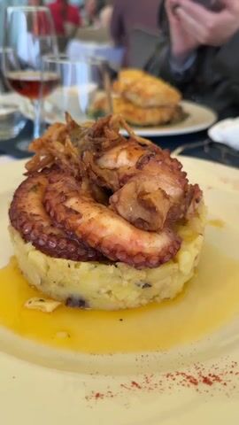 The video shows a plate of food consisting of a scoop of mashed potatoes topped with octopus and fried onions, drizzled with oil and sprinkled with paprika. A glass of red wine is visible in the foreground, and other plates of food and diners are visible in the background. The food is presented at an outdoor restaurant. A person is looking at their phone in the background.