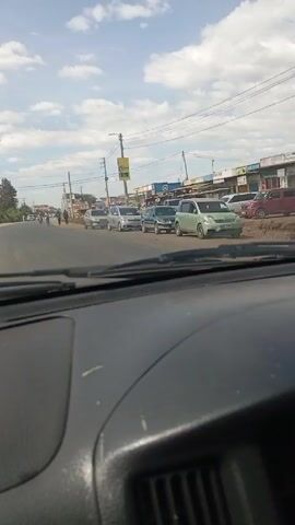 A car travels down a road. The road is flanked by a row of parked cars on the right. On the right side of the road are also several commercial buildings with storefronts. A motorbike passes the car going in the opposite direction. The motorbike is carrying a gas tank and other items. Several people walk along the road in the distance. Utility poles line the roadside. The sky is bright and cloudy.