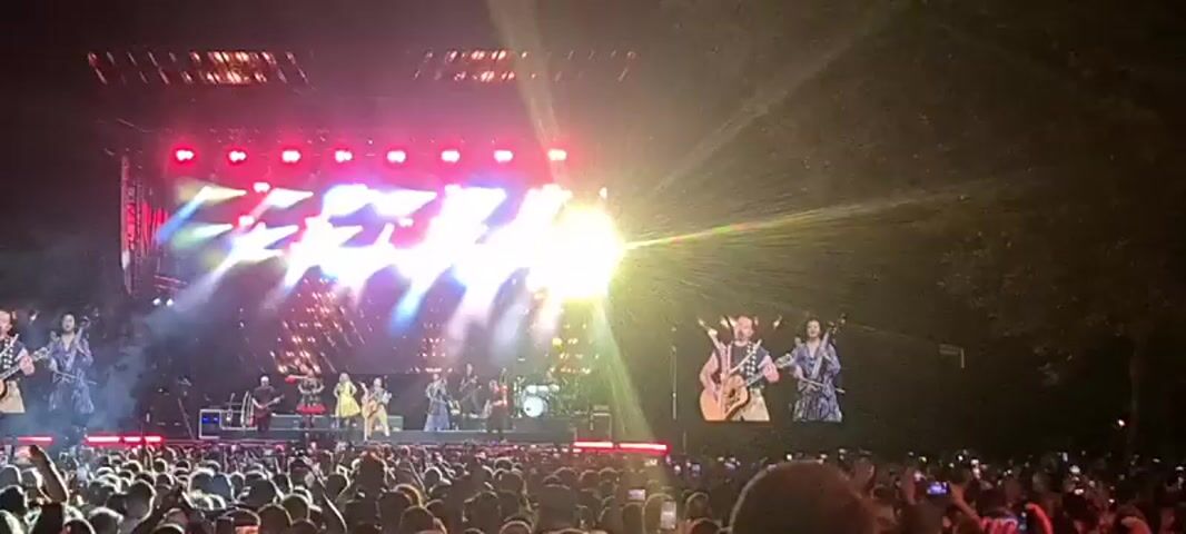 A large outdoor concert is taking place at night. A band performs on a brightly lit stage with a large screen behind them displaying images of the performers. A massive crowd of people is visible in the foreground, many holding up their phones to record the event. The atmosphere is energetic, with flashing lights and a sense of excitement.
