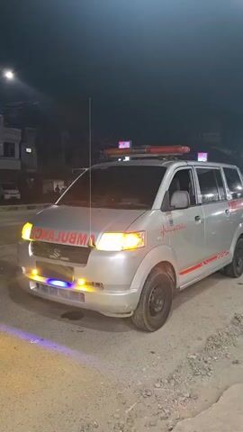 The video shows a silver ambulance with red and white markings. It is parked on a dirt road at night, with buildings and streetlights visible in the background. The ambulance has its emergency lights on, with red lights on the roof and blue and yellow lights on the front. The words "AMBULANCE" are visible in red letters on the front of the vehicle. Several people are visible near a building labeled "TERMINAL KOTA TUL".