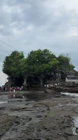 The video shows a temple situated on a rocky island, with many people gathered around it. The island is covered with green vegetation. The sky is overcast. The foreground is a wet, rocky surface. Some people are walking towards the temple, while others are standing or sitting around the base. The temple itself is a multi-tiered structure with trees growing on top. The island and temple appear to be surrounded by water.