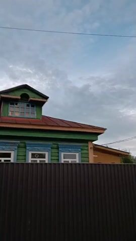 The video begins with a close-up of a wooden fence and a section of a house with green and blue trim and a brown roof. The camera then pans to the right, revealing a street with power lines overhead and a concrete pole with a streetlamp. Further down the street, more houses and fences are visible, some with red siding. The sky is cloudy and overcast. The video continues to pan right, showing more of the street and the houses lining it, with trees and foliage in the background. A car is parked on the side of the road. The camera eventually stops panning and the video ends with a view of the street, houses, and cloudy sky.