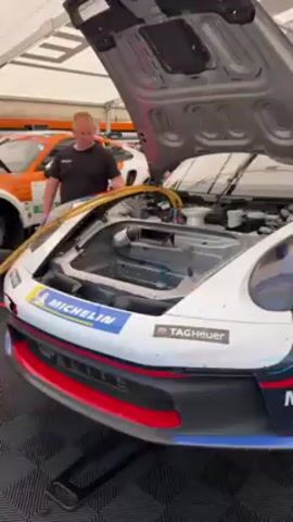 Here is a detailed description of the video:

The video shows a race car being refueled with the hood up. A mechanic is shown working on the car’s components. The car is white with Michelin and Tag Heuer stickers on the bumper. There are other mechanics nearby tending to other race cars. All of the cars are in a white garage or tent.