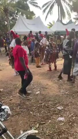 The video shows a cultural celebration, likely in Africa, where people in traditional costumes are dancing in an outdoor gathering. The costumes are colorful and elaborate, with some figures wearing masks. A man in a red shirt and black pants is dancing in the foreground. There are tents set up in the background. The ground is sandy and there are trees visible. A motorcycle is parked at the edge of the video. The atmosphere is lively, with music and movement.