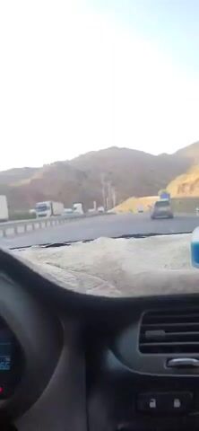 The video shows a view from inside a car looking out through the windshield at a highway. The car is moving, as indicated by the motion of the vehicles ahead and the blurred background. The highway passes through a mountainous, arid region. Several trucks and cars are visible on the highway. A car air freshener is placed on the dashboard. The driver's side dashboard with a digital display showing "46" is partially visible in the lower left corner. The sun appears to be shining brightly, suggesting it is daytime. A voiceover states, "You are in the radar zone. Camera in the back."