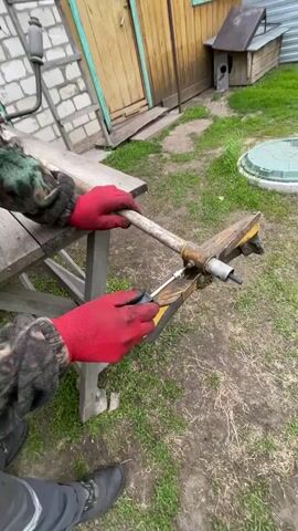A man in camouflage and red gloves is working on a piece of equipment on a workbench outside. The man is tightening a bolt on the piece of equipment using a wrench with a long handle. The equipment is yellow, rusty, and appears to be some type of tiller or cultivator attachment. The workbench is made of wood and has a trestle base. The ground is covered in grass and some dead leaves. Behind the workbench are a brick building with a wooden door, and a small dog house. There is also a gas powered machine in the background. The man is wearing glasses and a beanie. He is focused on his task.