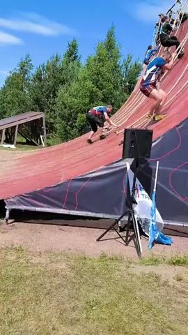 A low-angle, outdoor, medium shot captures participants in an obstacle course event scaling a large, curved red wall with ropes. The wall, which appears to be made of a flexible material, is tilted at a steep angle, requiring participants to use the ropes to ascend. A group of people are visible at the top of the wall, observing and possibly assisting. Some participants are already halfway up the wall, gripping the ropes and using their legs to push off the surface. Others are lower down, starting their ascent. The environment is bright and sunny, with a clear blue sky and some trees visible in the background. A speaker on a tripod stands at the base of the wall, suggesting that music or announcements are part of the event. Several flags are also present, indicating a sponsored event. The overall scene depicts a challenging physical activity within an organized competition.