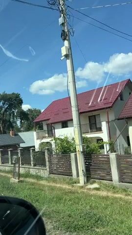 The video shows a view from a car driving past houses and trees under a blue sky with white clouds. In the foreground, the side mirror of a car is visible. On the right side of the road, there is a white house with a red roof and a fence. Further down the road, there is a sign that reads "HANUL DRAGUSEN PENSUNE TURISTICA". Trees, including pine trees, are abundant in the scene. There are also solar panels visible on one of the roofs. A truck with an open bed is parked on the left side of the road. There are no people visible in the video. The overall scene suggests a rural or suburban environment.