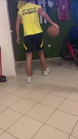 A man is filmed from behind, dribbling a basketball indoors. He wears a yellow t-shirt with "ФК МИНЬОР ЕЛШИЦА" in Cyrillic script on the back, black shorts with yellow trim, and white sneakers. The setting features a light-colored tiled floor and a dark green wall. On the wall, several sports jerseys are displayed, including a yellow one with "WINBET," a black and white one with "efbet," a red and white one with "A1," and a blue one. A red mountain bicycle is parked against the wall to the right. A broom is visible on the left side of the frame near a doorway. The man intermittently dribbles the basketball.