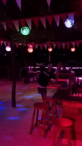 The video depicts an outdoor bar called "Ombak Bar," presumably at night, with festive lighting. A person in a dark t-shirt and shorts dances near the bar.  The bar is decorated with flags and spherical lights. Two individuals are visible behind the bar, possibly DJs. Surfboards are placed on either side of the bar, adding to the beach vibe. The ground appears to be sand. The scene is lit with a combination of red, blue, and white lights, creating a vibrant, club-like atmosphere.